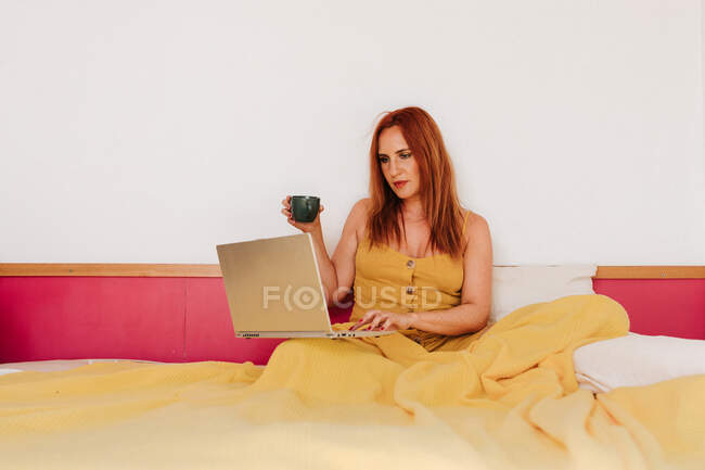Rothaarige Freiberuflerin in gelbem Kleid arbeitet mit Laptop, während sie Kaffee trinkt und auf dem Bett liegt — Stockfoto