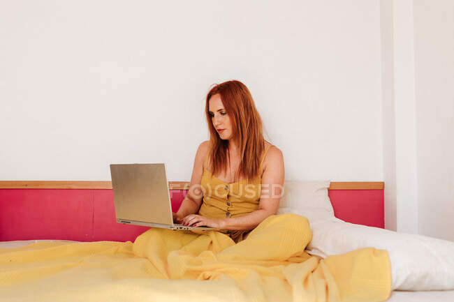 Rossa freelance femminile in lenzuola gialle che lavora con computer portatile sdraiato sul letto — Foto stock