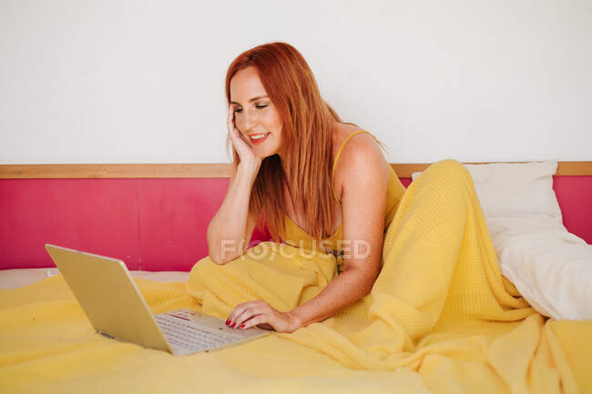 Рудий фрілансер жіночої статі в жовтих листах, що працюють з ноутбуком, лежачи на ліжку — стокове фото