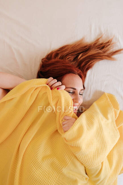 Vue du dessus rousse drôle de femme souriant tandis que sous couverture jaune à la maison avec les yeux fermés — Photo de stock