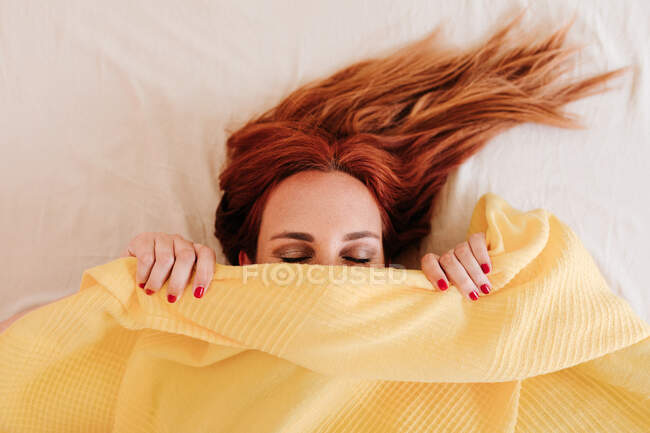 Vue du dessus rousse drôle de femme souriant tandis que sous couverture jaune à la maison avec les yeux fermés — Photo de stock