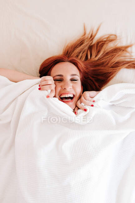 De dessus vue de dessus de rousse surprise femme drôle souriant tout en regardant sous la couverture blanche à la maison — Photo de stock