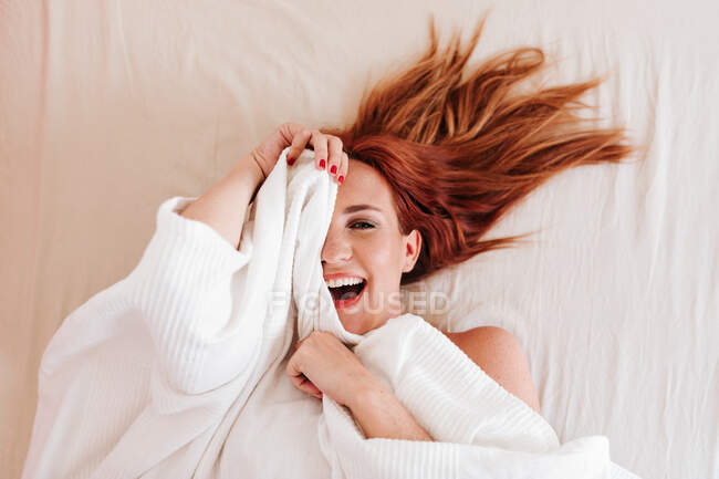 De dessus vue de dessus de rousse surprise femme drôle souriant tout en regardant sous la couverture blanche à la maison — Photo de stock