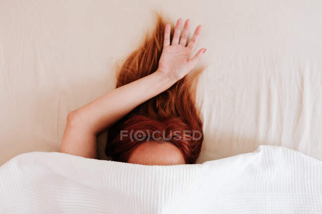 Visão superior do cabelo vermelho da senhora sem rosto com braço saliente ter relaxamento enquanto deitado na cama sob lençóis brancos — Fotografia de Stock