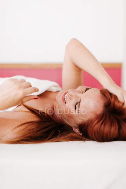 Веселая рыжая смешная женщина улыбается, лежа в постели с белым одеялом дома с закрытыми глазами — стоковое фото