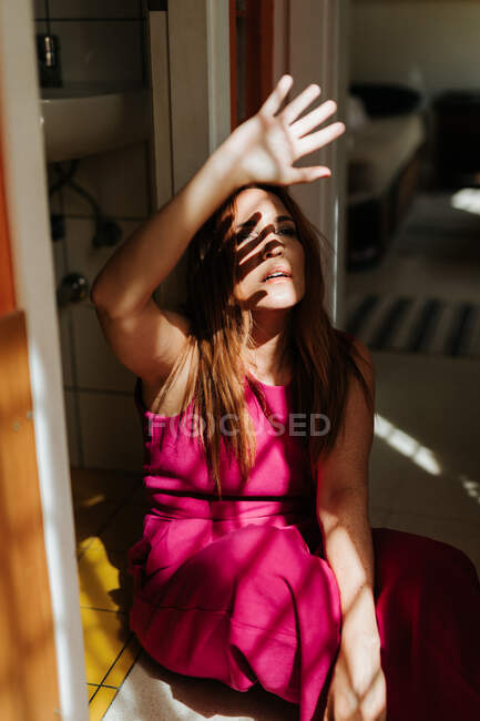 Ragionevole rossa donna in elegante abito rosa seduta con le gambe incrociate sul pavimento e guardando lontano con raggi di sole sul viso — Foto stock