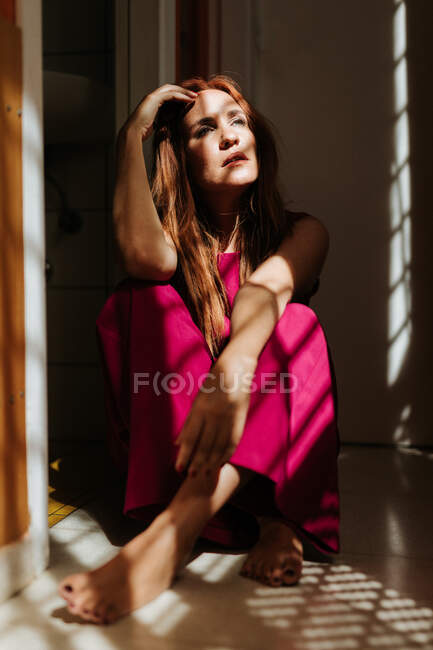 Mulher ruiva cuidadosa em vestido rosa elegante sentado com pernas cruzadas no chão e olhando para longe com raios de sol no rosto — Fotografia de Stock