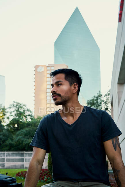 Jovem estudante hispânico masculino em roupas casuais elegantes sentado em cerca balançada e olhando para longe com o centro da cidade em segundo plano em Dallas, Texas — Fotografia de Stock