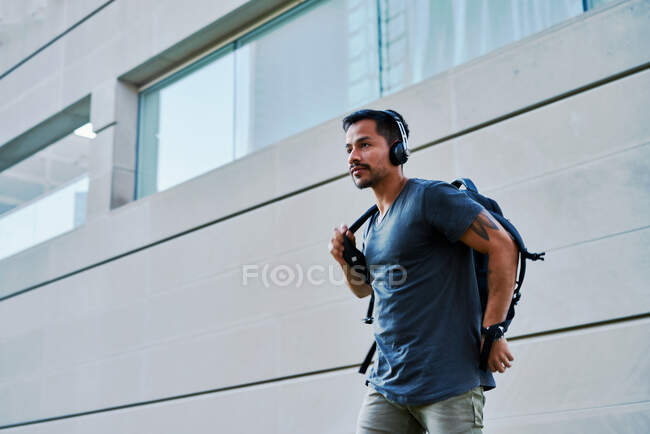 From below of Hispanic man in casual wear with headphones standing and putting on backpack at city street — Stock Photo
