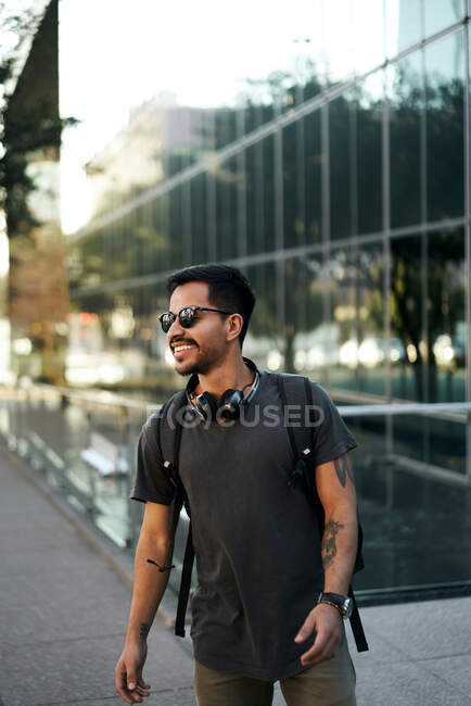 Joyeux homme hispanique barbu en tenue décontractée souriant et regardant loin avec des lunettes de soleil et des écouteurs à la mode sur le cou à la rue de la ville — Photo de stock