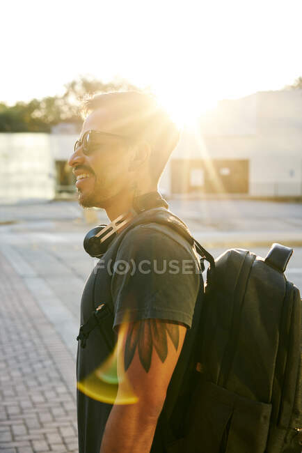 Hombre joven hispano en ropa casual y gafas de sol con estilo con auriculares y mochila mirando hacia la calle de la ciudad - foto de stock