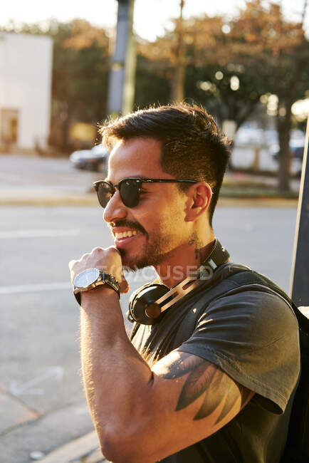 Молодий латиноамериканець у повсякденному одязі і стильних сонцезахисних окулярах з навушниками і рюкзаком, що дивляться на вулицю міста. — стокове фото