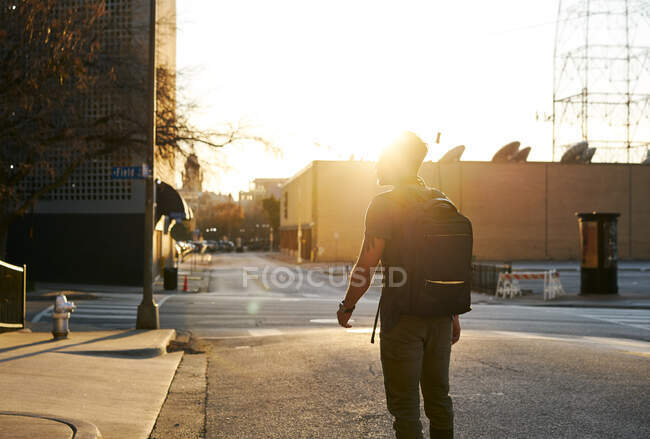 Вид ззаду на невпізнаваного чоловіка в повсякденному вбранні з рюкзаком, що йде по міській вулиці під час заходу сонця — стокове фото
