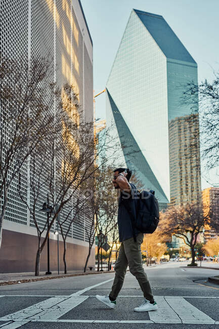 Viajante masculino hispânico em roupa casual e óculos de sol com mochila colocando fones de ouvido enquanto passeia no centro da cidade — Fotografia de Stock