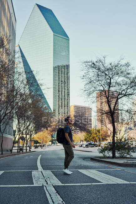 Feliz viajante hispânico masculino em roupa casual e óculos de sol com mochila com fones de ouvido enquanto passeia no centro da cidade — Fotografia de Stock