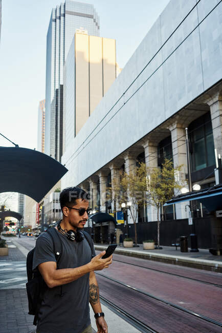 Сторона зору сучасного іспанського чоловіка туриста в повсякденному одязі і сонцезахисних окулярах з рюкзаком і навушниками за допомогою мобільного телефону на сучасній вулиці міста — стокове фото