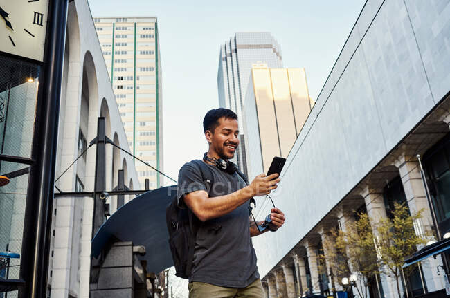 Turista masculino hispano contemporáneo en ropa casual y gafas de sol con mochila y auriculares usando teléfono móvil en la calle moderna de la ciudad - foto de stock