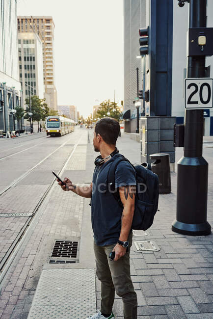 Anonymous Hispanic man in casual wear with backpack surfing on mobile phone while standing on platform at city street — Stock Photo