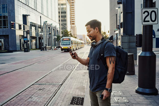 Латиноамериканець у повсякденному одязі з рюкзаковим серфінгом на мобільному телефоні, стоячи на платформі на вулиці міста — стокове фото