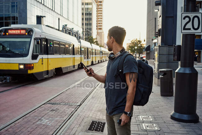 Анонімний іспанський чоловік у повсякденному одязі з рюкзаком серфінг на мобільному телефоні, стоячи на платформі на вулиці міста — стокове фото