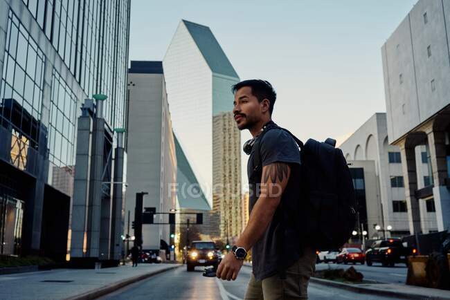 Vista lateral do viajante masculino hispânico em roupas casuais com mochila e fones de ouvido no pescoço andando ao longo da rua moderna da cidade — Fotografia de Stock