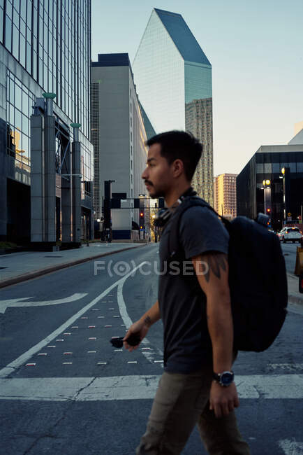 Vista lateral del viajero masculino hispano en ropa casual con mochila y auriculares en el cuello caminando por la calle moderna de la ciudad - foto de stock