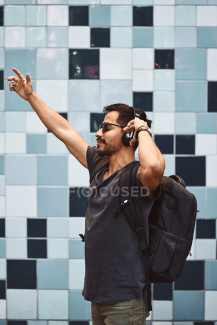 Homem hispânico feliz em roupas casuais e mochila com fones de ouvido em pé ouvindo música na rua da cidade — Fotografia de Stock