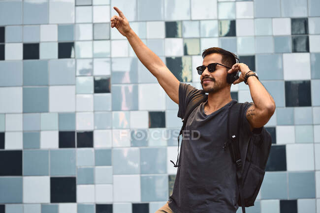 Hombre hispano feliz en ropa casual y mochila con auriculares de pie escuchando música en la calle de la ciudad - foto de stock