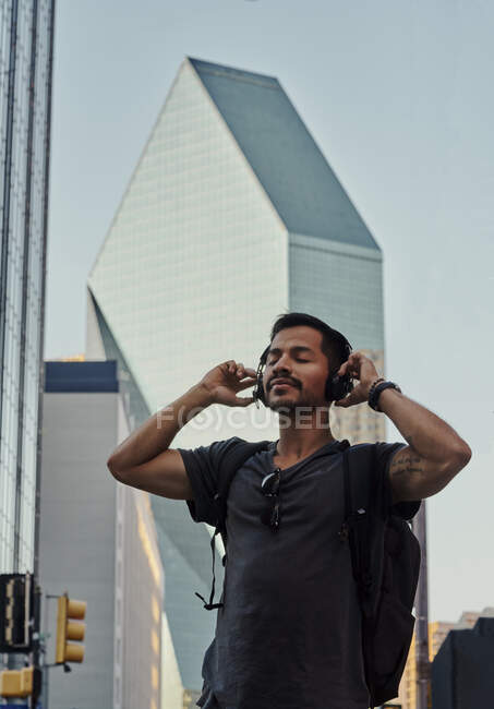 Happy hispanic man with closed eyes in casual wear and backpack with headphones standing listening to music at city street — Stock Photo