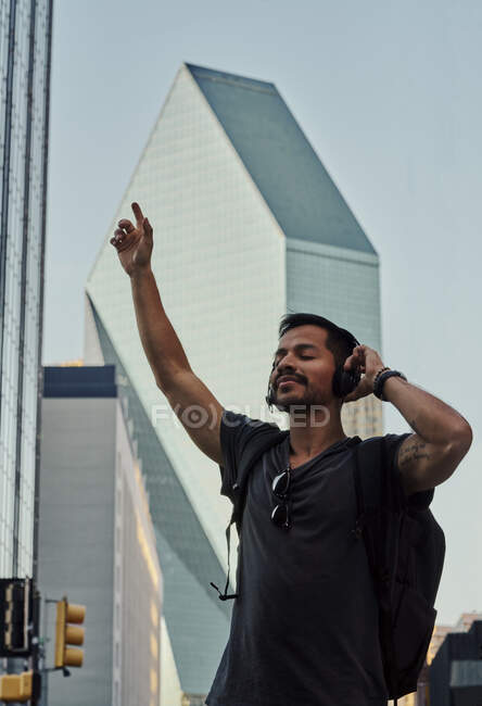 Hombre hispano feliz con los ojos cerrados en ropa casual y mochila con auriculares de pie escuchando música en la calle de la ciudad - foto de stock