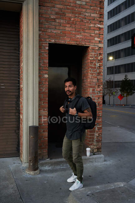 Side view of hispanic male traveler in casual outfit with backpack standing along empty city street with bricked building on background — Stock Photo