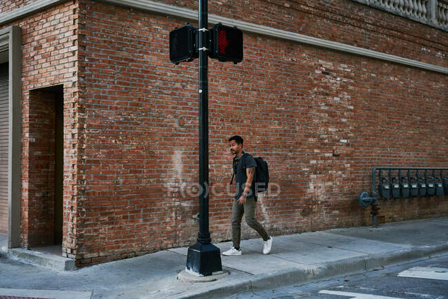 Estudiante masculino hispano en traje casual con mochila caminando por la calle vacía de la ciudad con edificio de ladrillo en el fondo - foto de stock