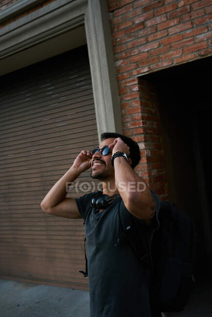 Вид сбоку на латиноамериканского путешественника в повседневной одежде и солнцезащитные очки стилиста с рюкзаком, стоящим вдоль пустой городской улицы с кирпичным зданием на заднем плане — стоковое фото