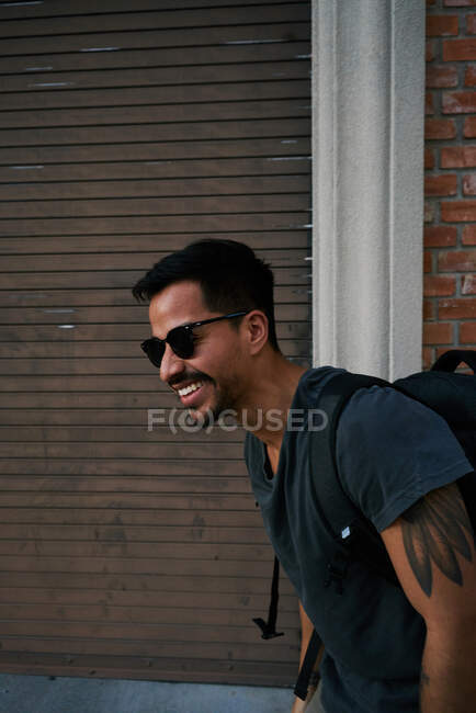 Side view of hispanic male traveler in casual outfit and stylist sunglasses with backpack standing along empty city street with bricked building on background — Stock Photo