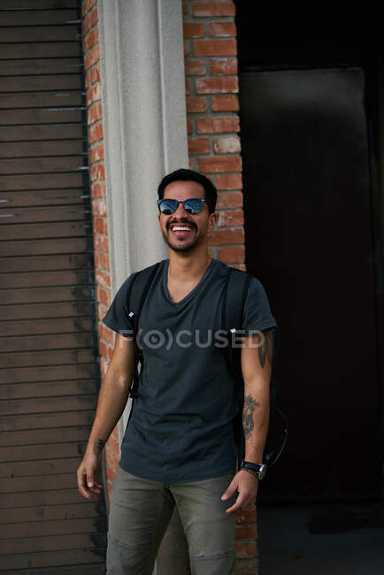 Hombre hispano viajero en traje casual y gafas de sol estilista con mochila de pie a lo largo de la calle vacía de la ciudad con el edificio de ladrillo en el fondo - foto de stock
