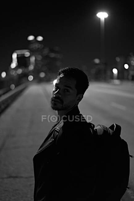 Vista lateral do homem hispânico na moda em jaqueta de couro preto casual com mochila olhando para a câmera com cidade noturna no fundo embaçado — Fotografia de Stock