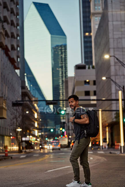 Vista lateral do viajante masculino hispânico satisfeito com mochila em roupa casual rindo enquanto passeia no centro moderno — Fotografia de Stock