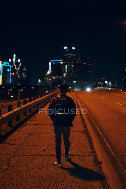 Back view of anonymous man in casual black leather jacket with backpack walking at night city on blurred background — Stock Photo