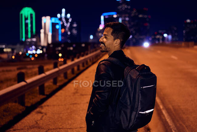 Vista lateral do homem hispânico na moda em jaqueta de couro preto casual com mochila olhando para longe com a cidade noturna no fundo embaçado — Fotografia de Stock