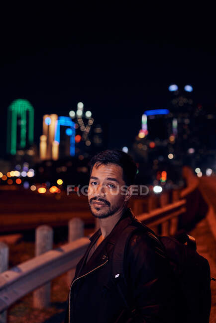 Side view of fashionable Hispanic man in casual black leather jacket with backpack looking at camera with night city on blurred background — Stock Photo
