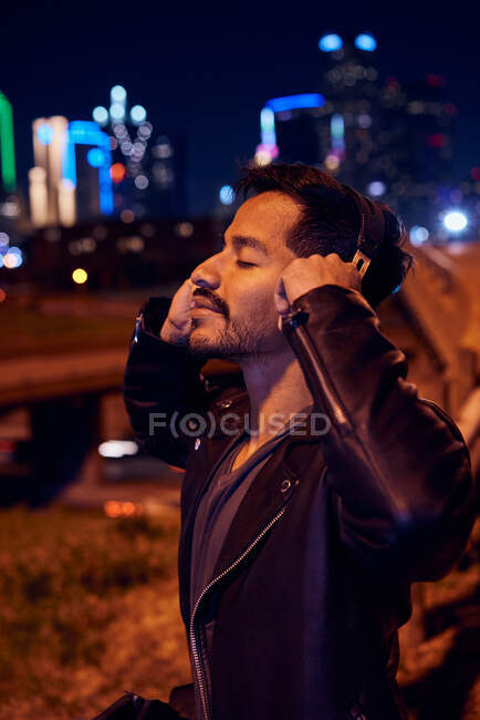 Satisfied Hispanic man in stylish leather black jacket smiling while listening music with headphones at modern city street — Stock Photo