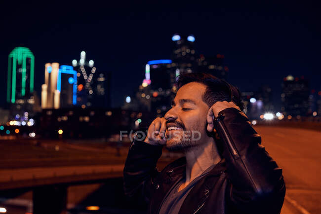 Hombre hispano satisfecho en elegante chaqueta de cuero negro sonriendo mientras escucha música con auriculares en la calle moderna de la ciudad - foto de stock