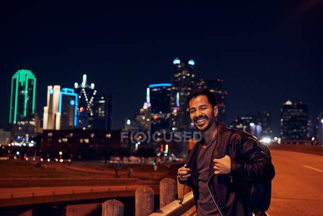 Homme hispanique à la mode en veste décontractée en cuir noir avec sac à dos regardant la caméra avec ville nocturne sur fond flou — Photo de stock