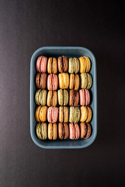 Macaroons saborosos coloridos exibidos dentro do recipiente azul no fundo preto — Fotografia de Stock
