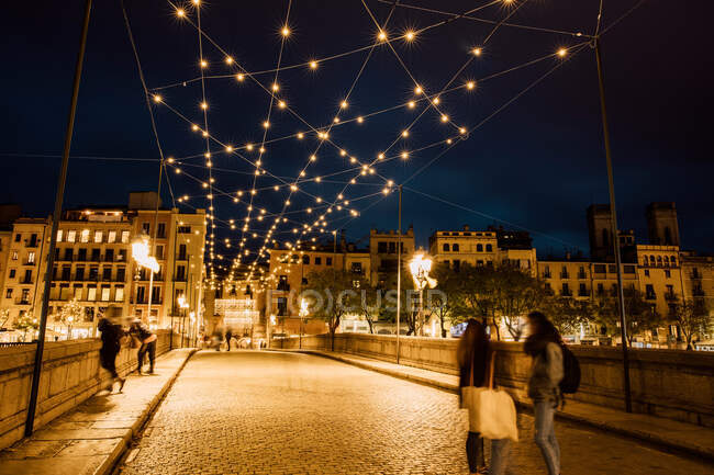 Personas relajadas apoyadas en valla y caminando sobre puente bajo iluminación en Girona, Cataluña, España - foto de stock