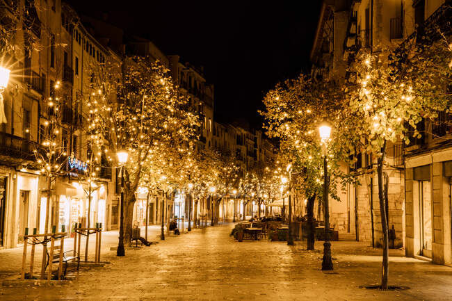 Вечерний пейзаж аллеи в теплом ярком свете от освещения и фонарей вдоль улицы, украшенной деревом в центре Жироны, Каталония, Испания — стоковое фото