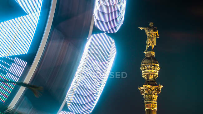 Colorata ruota panoramica illuminata nella vista notturna del paesaggio cittadino e una statua monumentale — Foto stock