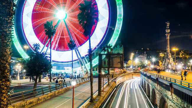 Красочный пейзаж ночного города с видом на дорогу и освещенное колесо обозрения и улицу, украшенную пальмами — стоковое фото