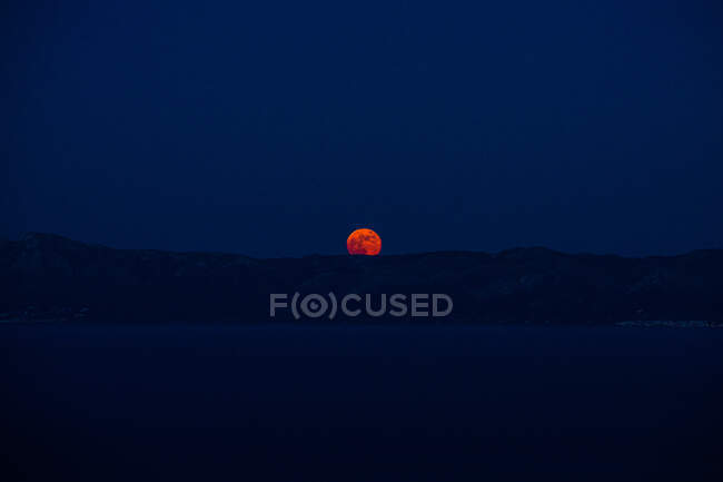 Dark landscape with orange sun hiding in row of mountain above calm water — Stock Photo