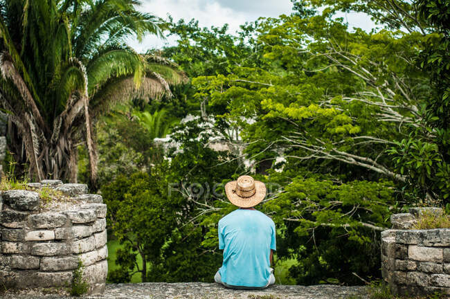 Vista posteriore del maschio senza volto in abbigliamento casual e cappello esplorare la natura seduto sul tetto del vecchio edificio — Foto stock